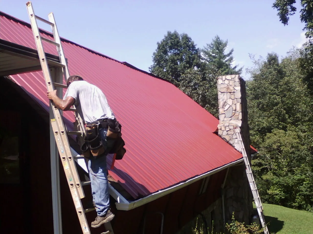 Roofer on a ladder beside a red metal roof and stone chimney — Soffit & Fascia Repair by Roofing Brothers in Jay Em